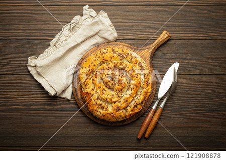 Burek made of phyllo dough with filling on cutting board, dark brown wooden rustic background top view. Traditional savoury spiral pie of Balkans, Middle East and Central Asia 121908878