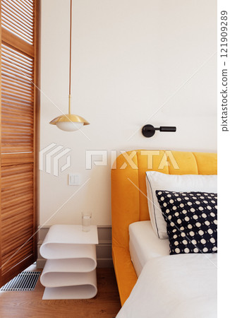 Close-up of a stylish modern bedside table with a glass of water. A cozy bed with contrasting pillows and a yellow upholstered headboard. 121909289