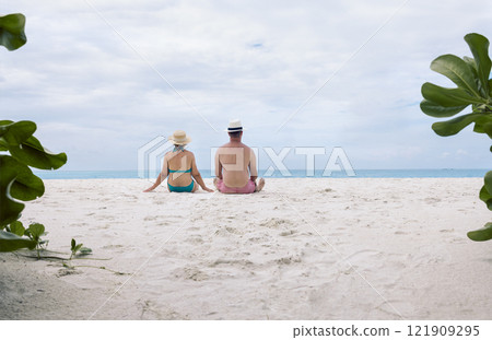 A young married couple is sitting on the sand on the beach and enjoying the beautiful seascape. A man and his wife in swimsuits on the ocean shore. The view from the back. 121909295