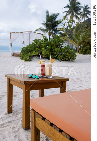 Two cocktails with slices of fruit are on a wooden table on the beach. Delicious chilled drinks on the background of tropical trees and the ocean. Two cocktails with slices of fruit are on a wooden table on the beach. Delicious chilled drinks on the background of tropical trees and the ocean. 121909296