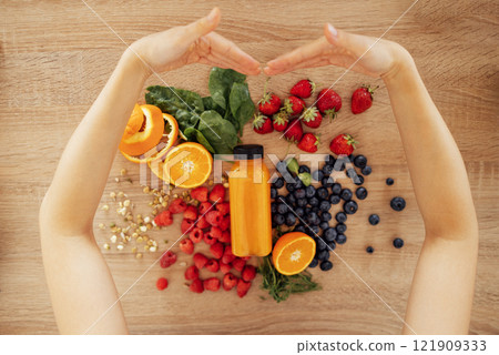 Female hands embracing citrus fruits, berries, greens, sprouted beans and bottle of orange juice on the background of a wooden table. 121909333