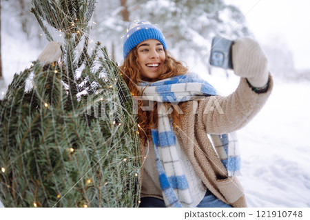 A young woman with a phone and a Christmas tree with garlands enjoys the beautiful winter weather. 121910748