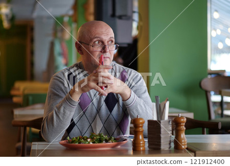 Senior man in glasses sits at table in restaurant and holds red smartphone Senior man in glasses sits at table in restaurant and holds red smartphone 121912400