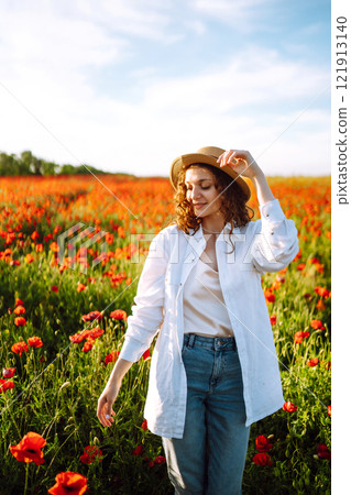 Young girl in hat posing in the poppy field. Portrait of curly girl walking in poppy field at sunset. Summertime. 121913140