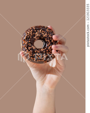 Hand holding tglazed chocolate donut on mocha brown background. Hand holding tglazed chocolate donut on mocha brown background. 121913335