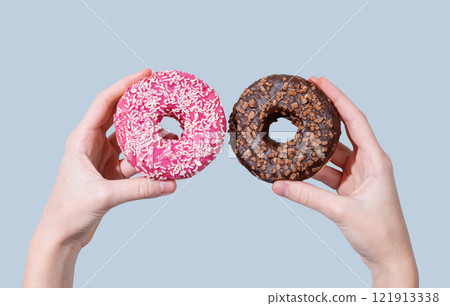 Hands holding two glazed chocolate and pink donuts on blue background. Hands holding two glazed chocolate and pink donuts on blue background. 121913338