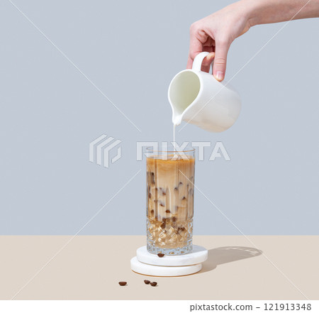 Cold latte coffee in a tall glass with hand pouring alternative milk from a milk jug  121913348