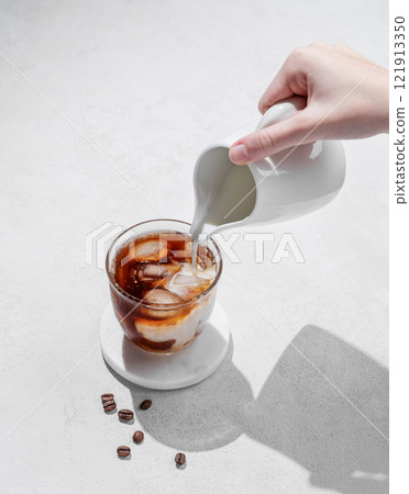 Cold latte coffee in a glass with hand pouring alternative milk from a milk jug on light background Cold latte coffee in a glass with hand pouring alternative milk from a milk jug on light background 121913350