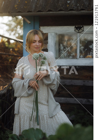 A rural woman stands in a garden with wild flowers against the backdrop of an old village house A rural woman stands in a garden with wild flowers against the backdrop of an old village house 121913761
