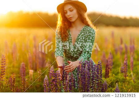 Close-up of young woman in hat and curly hair touching lupins in field at sunset. Fashion and nature 121913915