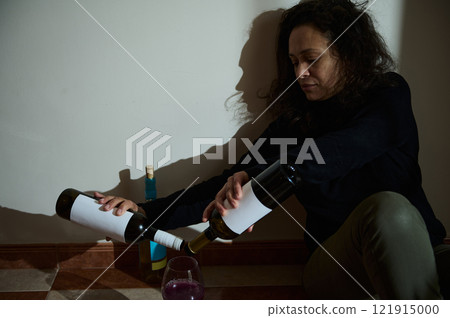 Woman sitting with wine bottles displaying emotional distress under dim lighting Woman sitting with wine bottles displaying emotional distress under dim lighting 121915000