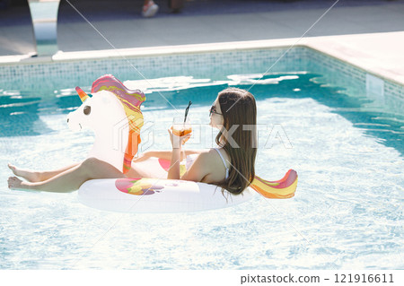 Young girl on a unicorn swim ring in the pool. Girl wearing white swimwear and a sunglasses. Brunette girl relaxing in the swimming pool drinking a cocktail. Young girl on a unicorn swim ring in the pool. Girl wearing white swimwear and a sunglasses. Brunette girl relaxing in the swimming pool drinking a cocktail. 121916611