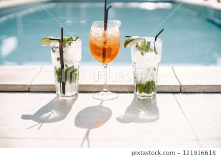 Cropped photo of glasses with orange beverage and two mohitos. Three cocktails standing near swimming pool. Focus on a drinks in glasses with a straw outdoors. 121916618