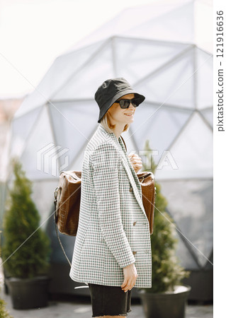 Outdoor portrait of a young beautiful fashionable woman walking on the street, holding big brown leather bag. Blonde girl wearing sunglasses and black hat. There are round glass building behind. 121916636