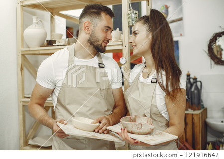 Mutual creative work. Young beautiful couple in casual clothes and aprons. People holds ceramic dishes. 121916642
