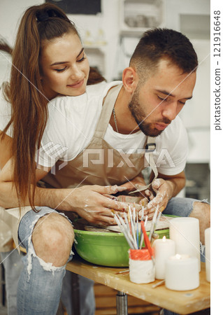 Mutual creative work. Young beautiful couple in casual clothes and aprons. People creating a bowl on a pottery wheel in a clay studio. Mutual creative work. Young beautiful couple in casual clothes and aprons. People creating a bowl on a pottery wheel in a clay studio. 121916648
