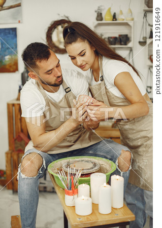 Mutual creative work. Young beautiful couple in casual clothes and aprons. People creating a bowl on a pottery wheel in a clay studio. Mutual creative work. Young beautiful couple in casual clothes and aprons. People creating a bowl on a pottery wheel in a clay studio. 121916649
