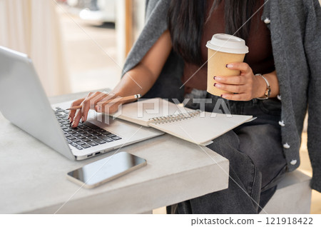 A close-up of a woman works remotely from a coffee shop, typing on a laptop keyboard. 121918422