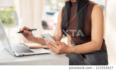 A woman works remotely from a coffee shop, using her smartphone while working on her laptop. 121918427