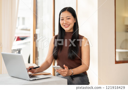 A beautiful Asian woman working remotely from a coffee shop in the morning, working on her laptop. 121918429