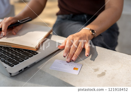 A close-up of a woman's hand holds a credit card above a table while using her laptop. A close-up of a woman's hand holds a credit card above a table while using her laptop. 121918452