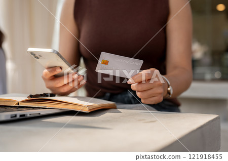 A close-up of a woman's hand holds a credit card and a smartphone above a table, shopping online. A close-up of a woman's hand holds a credit card and a smartphone above a table, shopping online. 121918455