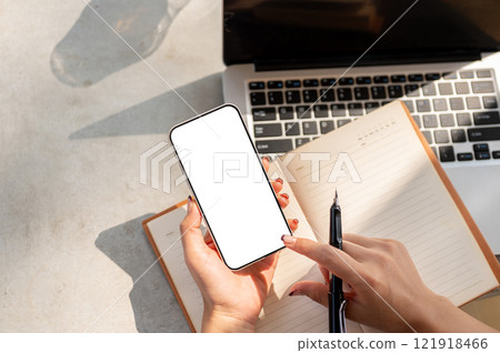 Top view of a woman's hand holding a smartphone with a white screen mockup above a desk. Top view of a woman's hand holding a smartphone with a white screen mockup above a desk. 121918466