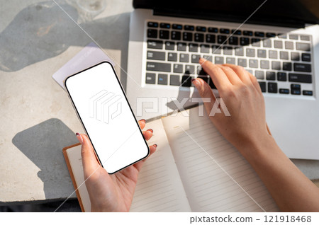 Top view of a woman's hand holding a smartphone with a white screen mockup above a desk. 121918468