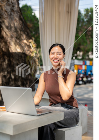 Attractive Asian businesswoman talks on the phone with her client while working remotely from a cafe Attractive Asian businesswoman talks on the phone with her client while working remotely from a cafe 121918474