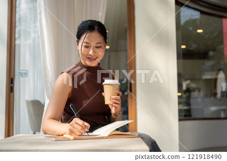 A beautiful, positive Asian woman writes her diary while enjoying a hot drink at a cafe. 121918490