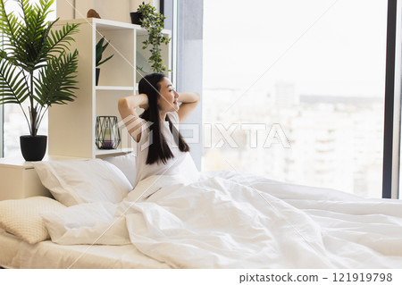Asian woman in bright bedroom stretches in bed surrounded by plants. Morning light fills room, creating serene atmosphere. Relaxation and comfort emphasized in modern living space. Asian woman in bright bedroom stretches in bed surrounded by plants. Morning light fills room, creating serene atmosphere. Relaxation and comfort emphasized in modern living space. 121919798