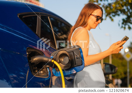 Woman in a white dress stands beside an electric car, checking her smartphone while it charges. Woman in a white dress stands beside an electric car, checking her smartphone while it charges. 121920674