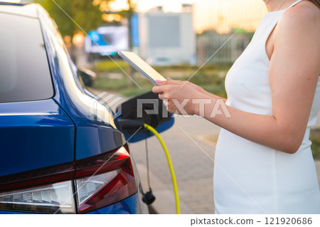 Pregnant woman in white dress charging an electric car and using her mobile phone to pay Pregnant woman in white dress charging an electric car and using her mobile phone to pay 121920686