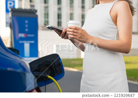 Woman in a white dress charges her electric vehicle, holding coffee and a smartphone in hand 121920687