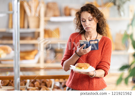 European female customer with phone takes pictures of burger croissant in disposable plate dish European female customer with phone takes pictures of burger croissant in disposable plate dish 121921562