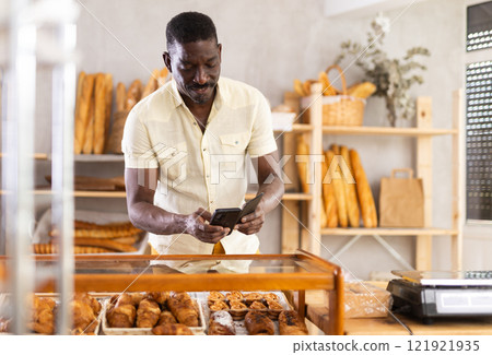 African male customer with phone takes pictures of buns in bakery window 121921935