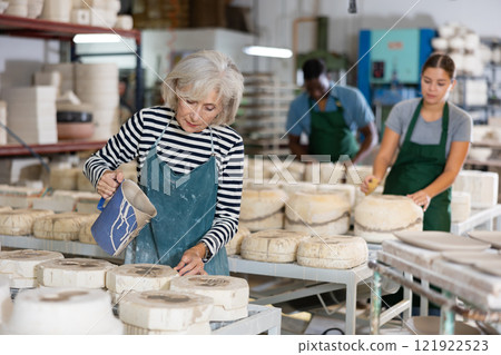 Mature worker pours slip mass from jug into plaster mold in pottery factory 121922523