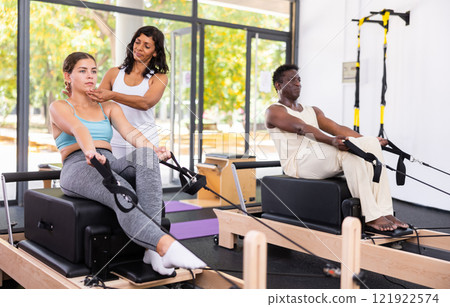 Latin female instructor helping positive girl practicing pilates on reformer Latin female instructor helping positive girl practicing pilates on reformer 121922574