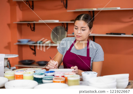 Woman potter with paintbrush glazing, painting on plate in workshop, working in pottery 121922667