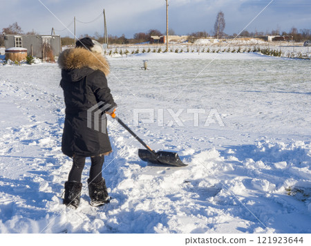 Woman shoveling snow, bundled in winter attire, enjoying bright sunlight across snow-covered landscape 121923644