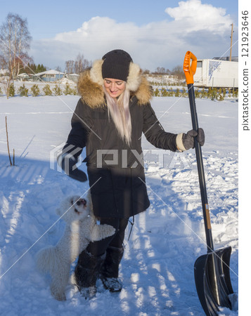 Cheerful woman shoveling snow with energetic white dog playing nearby, bright winter sunlight illuminating snowy backyard landscape 121923646