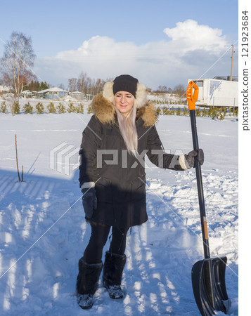 Woman wearing winter attire cheerfully clearing snow, enjoying bright sunlight across snowy landscape with infectious smile and energetic movement 121923684