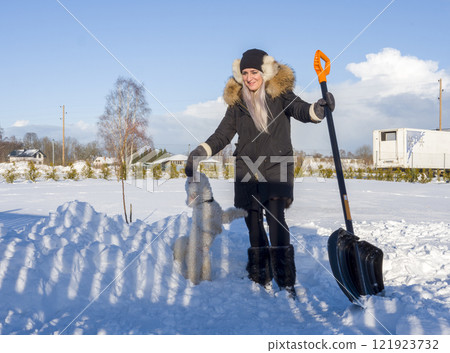 Woman shoveling snow, playing with white dog in sunny winter landscape, bundled in cold-weather gear 121923732