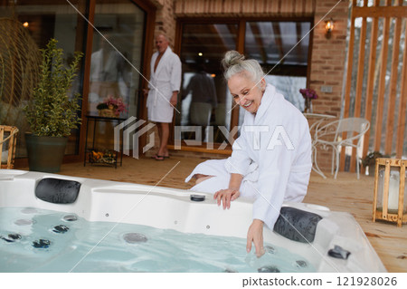 Older woman checking water temperature in hot tub with hand. Romantic wellness getaway for senior couple. Valentine's weekend. Older woman checking water temperature in hot tub with hand. Romantic wellness getaway for senior couple. Valentine's weekend. 121928026