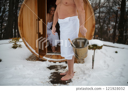 Older man entering small sauna, carrying bucket of water to create steam. Winter Spa weekend getaway for elderly people. Older man entering small sauna, carrying bucket of water to create steam. Winter Spa weekend getaway for elderly people. 121928032