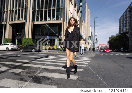 Stylish Young Fashion Woman With Long Hair Walking Across Pedestrian Crossing In City, Wearing Black Mini Dress And Boots, Trendy Urban Street Style Model Stylish Young Fashion Woman With Long Hair Walking Across Pedestrian Crossing In City, Wearing Black Mini Dress And Boots, Trendy Urban Street Style Model 121929514
