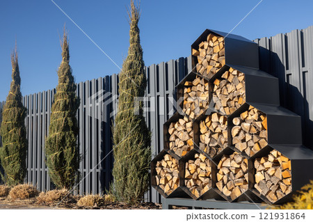 A row of tall thuja trees wrapped in protective twine stands next to a modern, geometric firewood storage rack filled with neatly stacked logs, against a grey metal fence and a clear blue sky. A row of tall thuja trees wrapped in protective twine stands next to a modern, geometric firewood storage rack filled with neatly stacked logs, against a grey metal fence and a clear blue sky. 121931864