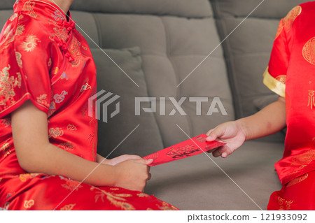 A happy sibling duo, dressed in red cheongsam qipao, exchange monetary gifts in hongbao envelopes at home. The living room scene captures the warmth and tradition of Chinese New Year 121933092