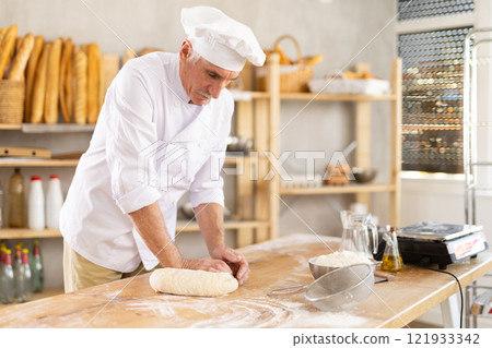 Elderly male baker preparing raw yeast dough on kitchen table in bakery 121933342