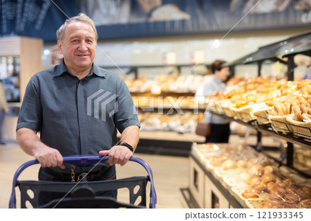 Elderly male shopper with a shopping cart chooses groceries in supermarket 121933345